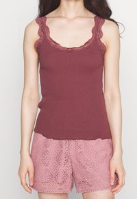 Donna che indossa un top senza maniche a coste color bordeaux con bordi in pizzo e pantaloni corti in pizzo rosa, in piedi contro uno sfondo neutro.