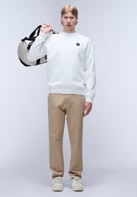 Felpa bianca con polsini a costine, pantaloni beige e sneakers bianchi. Tiene una borsa a tracolla beige con maniglie scure. Design semplice e moderno.