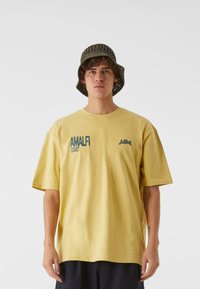 Gul bomulls T-skjorte med korte ermer, med "AMALFI COAST" trykt i grønt. Brukt med en teksturert olivenfarget bøttehatt.