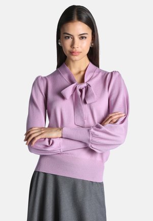 Femme aux cheveux longs et raides portant un pull violet clair avec un détail noeud papillon et une jupe grise, les bras croisés, debout devant un fond uni.