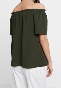 Vihreä olkapäältä avoin paita elastisella kauluksella, lyhyet hihat ja rento istuvuus. Silkkinen kangas laskeutuu pehmeästi, yhdistettynä vaaleisiin housuihin.