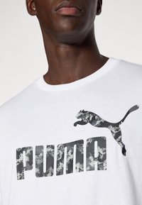 Balta medvilninė marškinėliai su dideliu pilku kamufliažo "PUMA" logotipu ir bėgančio puma grafika atitinkančiomis spalvomis. Trumpomis rankovėmis ir apvaliu iškirpimu.