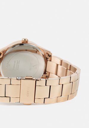 Náramek hodinek z růžově zlaceného kovu s přezkou, na které je vyryt nápis "GUESS", zobrazující zadní stranu pouzdra hodinek a korunku na bílém pozadí.