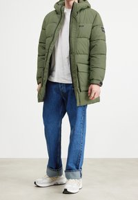 En person iført en grøn puffjakke, hvid t-shirt, opstillede blå jeans og hvide sneakers står på et ensfarvet gulv med en hvid baggrund.