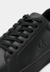 Zwart leren sneaker met platte zwarte veters, ronde neus, gestructureerde rubberen zool en een gegraveerd CK-logo aan de zijkant. Glad afwerking.