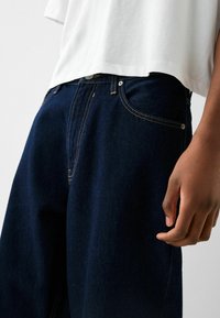 Tmavě modré džíny z denimu se zlatým prošíváním, designem s pěti kapsami a zapínáním na knoflík, s uvolněným střihem a hladkou texturou.