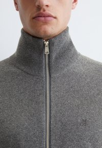 Gray zip-up sweater with a ribbed collar, featuring a silver zipper and subtle logo detail on the chest. Soft fabric texture is visible.