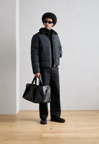 Polstret sort jakke med shearling krave, sort turtleneck, mørke bukser og en sort læder duffel taske. Glansfulde sorte sko.