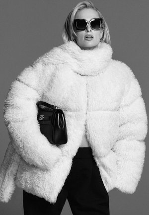 White fluffy oversized puffer jacket with a high collar, paired with black sunglasses and a small black handbag. Black pants complete the look.
