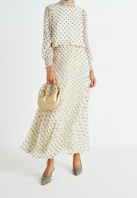 Abito midi a pois in crema e marrone, con gonna plissettata, scollatura alta e maniche lunghe. Abbinato a una borsa dorata e tacchi metallici.