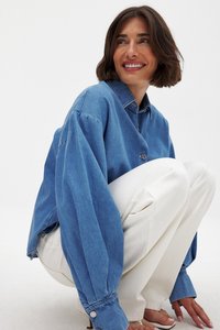 Helle blaue Jeansjacke mit durchgeknöpftem Frontverschluss, übergroßen Ärmeln und klassischem Kragen; kombiniert mit weißen, lockeren Hosen.