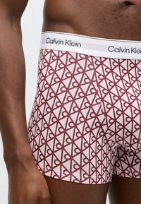 Ανδρικό εσώρουχο Calvin Klein τύπου boxer με λευκή μέση και γεωμετρικό μοτίβο κόκκινης καρδιάς σε φωτεινό φόντο, κατασκευασμένο από μαλακό βαμβακερό ύφασμα.