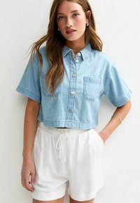 Camisa de mezclilla azul claro, corta y de manga corta, con un bolsillo frontal, combinada con pantalones cortos blancos con cordón. Textura suave y corte casual.