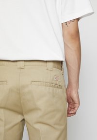 Beige chinos med en glatt tekstur, to baklommer og en liten merkepute på høyre lomme, kombinert med en hvit t-skjorte.