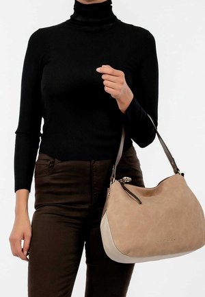 Person wearing black turtleneck and dark pants holds a beige leather handbag with dark shoulder strap on white background.