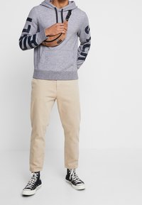 Grå hoodie med marinblå accenter och stort logotyp på ärmarna, tillsammans med beige chinos och svarta högstövlar. Innehåller en framficka och en justerbar axelväska.