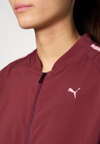 Burgundy urheilu takki, jossa on vetoketjufrontti, jossa on vaaleanpunainen Puma-logo ja vaaleanpunaiset raidat hartioissa, valmistettu sileästä kankaasta.