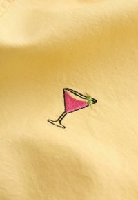 Tissu jaune avec un verre à cocktail rose brodé avec une garniture verte sur le bord.