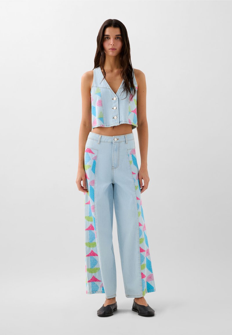 Vest scurt din denim bleu deschis cu nasturi argintii. Blugi largi asortați cu modele geometrice colorate în roz, albastru și verde.