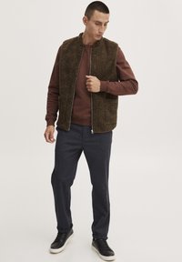 Brun fleece vest med lynlås foran og sidelommer, lagd over en brun sweatshirt, parret med mørkegrå strukturerede bukser og sorte sneakers.