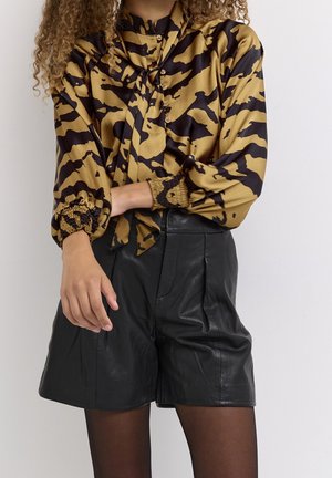 Mujer con una blusa estampada en dorado y negro con mangas abullonadas y pantalones cortos de cuero negros de cintura alta sobre un fondo liso.