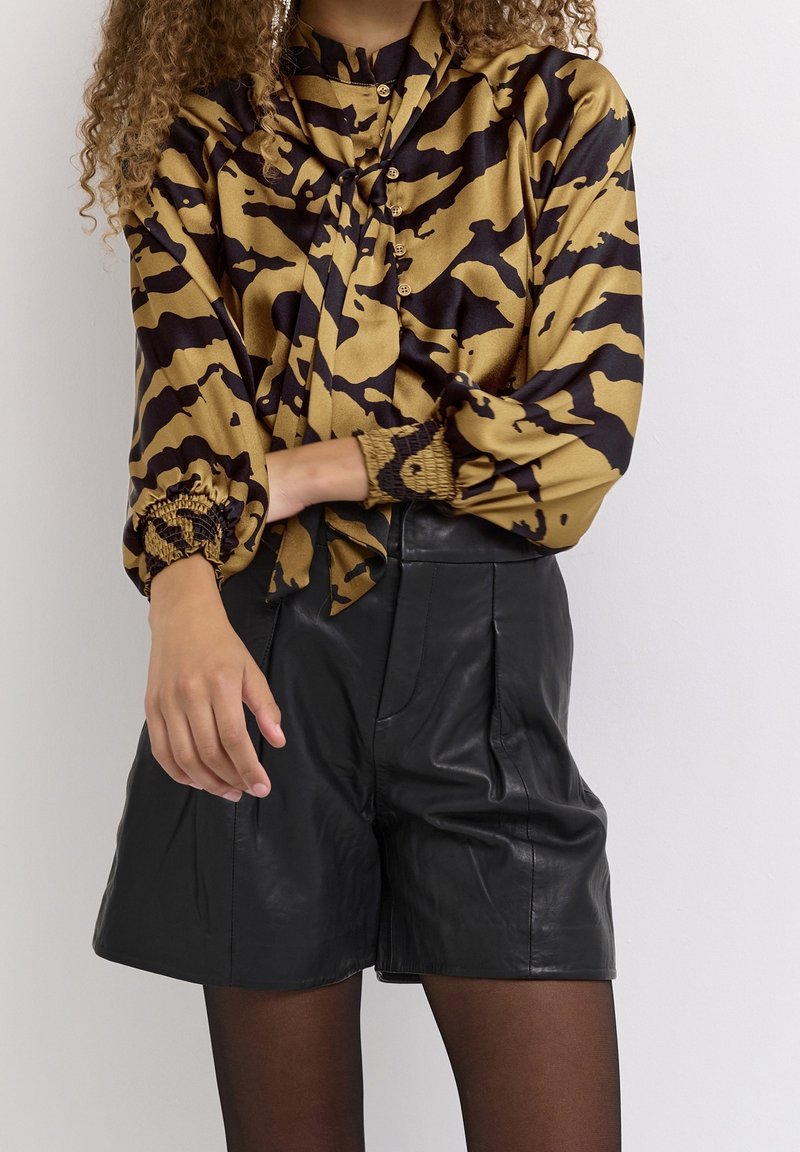 Femme portant un chemisier à motif doré et noir avec des manches bouffantes et un short en cuir noir taille haute sur un fond uni.