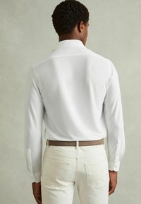 Cămașă albă cu mânecă lungă și manșete cu nasturi, purtată cu pantaloni de culoare deschisă și o curea maro. Textură netedă, guler clasic și design simplu.