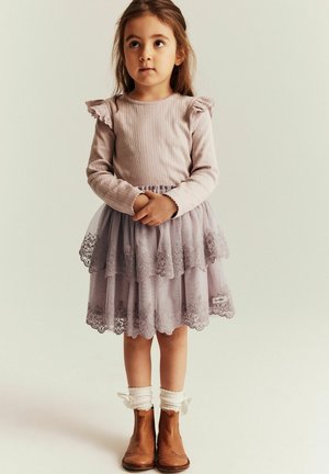 Jong meisje met lang haar, gekleed in een roze top met lange mouwen, een gelaagde lavendelkleurige kanten rok, witte sokken met strikjes en bruine enkellaarsjes.