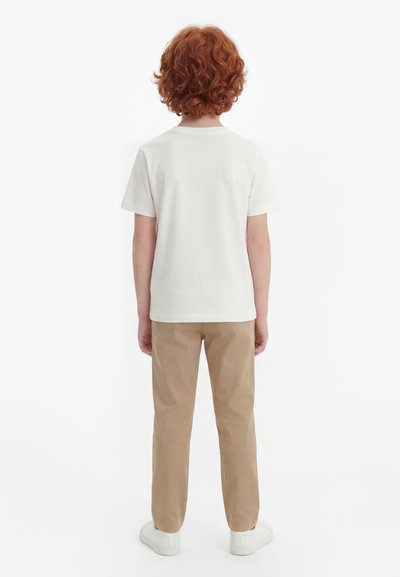 Dziecko z kręconymi rudymi włosami stoi odwrócone tyłem, ubrane w gładką białą koszulkę z krótkim rękawem, beżowe spodnie i białe trampki na białym tle.