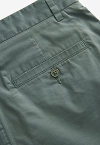 Shorts di tessuto verde presentano una tasca posteriore con un singolo bottone cucito, una texture liscia e cuciture ordinate lungo le cuciture.