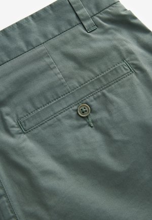 Shorts di tessuto verde presentano una tasca posteriore con un singolo bottone cucito, una texture liscia e cuciture ordinate lungo le cuciture.