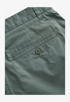 Zelené látkové šortky majú zadné vrecko s jedným prišitým gombíkom, hladkú textúru a úhľadné stehy pozdĺž švov.
