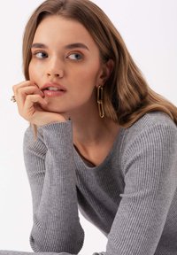 Szary sweter z prążkowanego materiału z okrągłym dekoltem. Modelka nosi duże złote kolczyki w kształcie spinaczy. Włosy są falowane, opadają na twarz i ramiona.