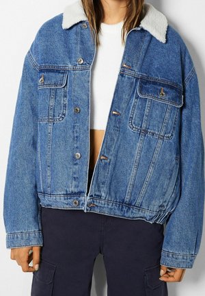 Jeansjacke mit weißem Fleecekragen, Druckknöpfen und Brusttaschen, getragen über einem weißen Oberteil und dunkler Hose.