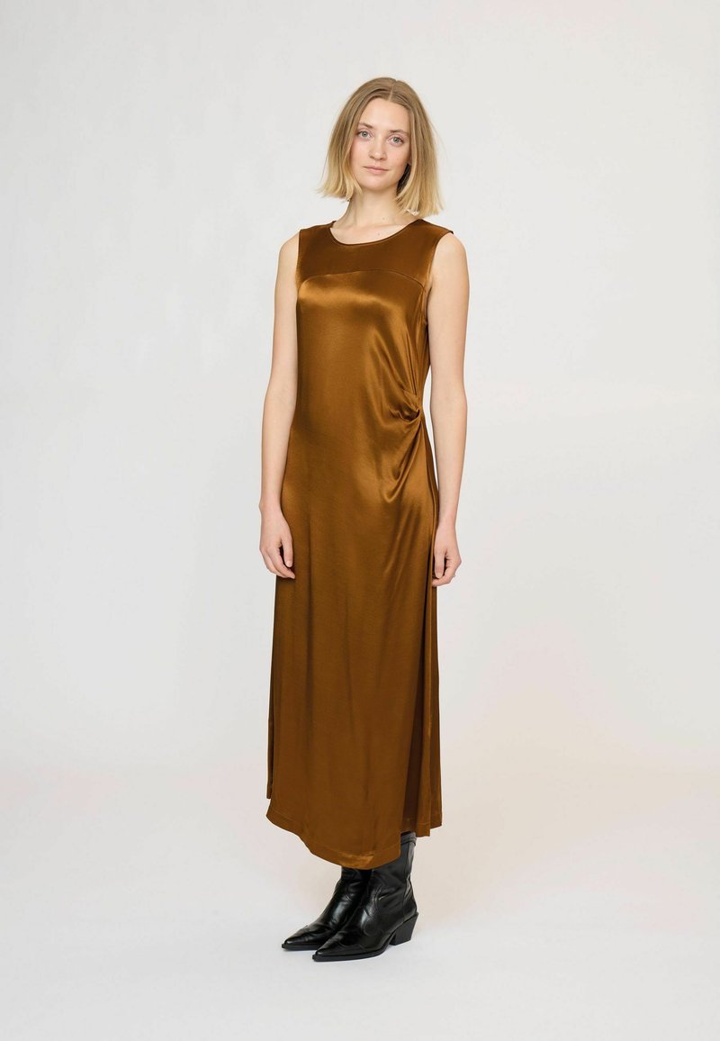 Femme aux cheveux blonds portant une longue robe marron en satin sans manches et des bottines noires, debout devant un fond blanc.