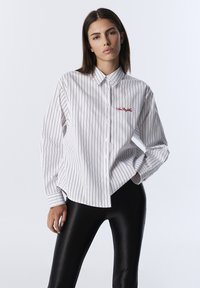 Camisa blanca de botones con rayas verticales negras, con un logo bordado en rojo. Mangas largas y un corte holgado. Combinada con leggings negros brillantes.