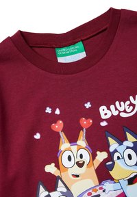 Maglietta color marrone rossiccio con personaggi animati di Bluey, tra cui un cane arancione con antenne a forma di cuore, che giocano e sorridono, della United Colors of Benetton.
