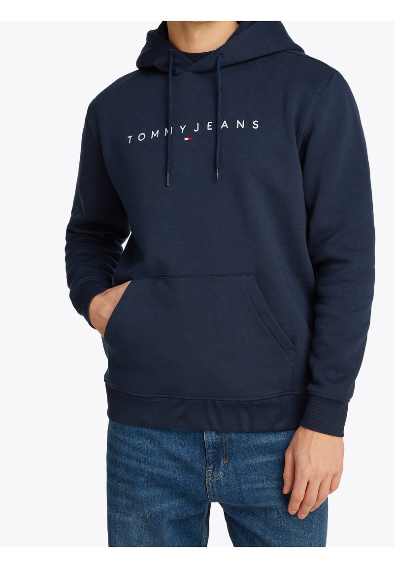 Mies, joka on pukeutunut tummansiniseen huppariin, jossa on "TOMMY JEANS" -logo ja lippu, sinisiin farkkuihin ja yksi käsi etutaskussa.