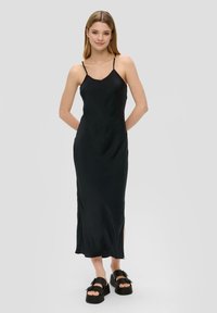 Schwarzes, ärmelloses Maxikleid mit glatter Textur, das einen dezenten V-Ausschnitt und seitliche Schlitzdetails aufweist. Kombiniert mit klobigen schwarzen Plateausandalen.
