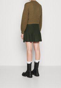 Suéter de punto verde oliva con un patrón texturizado, de longitud corta, combinado con una falda de campana verde oscuro y botas negras hasta el tobillo con calcetines blancos.