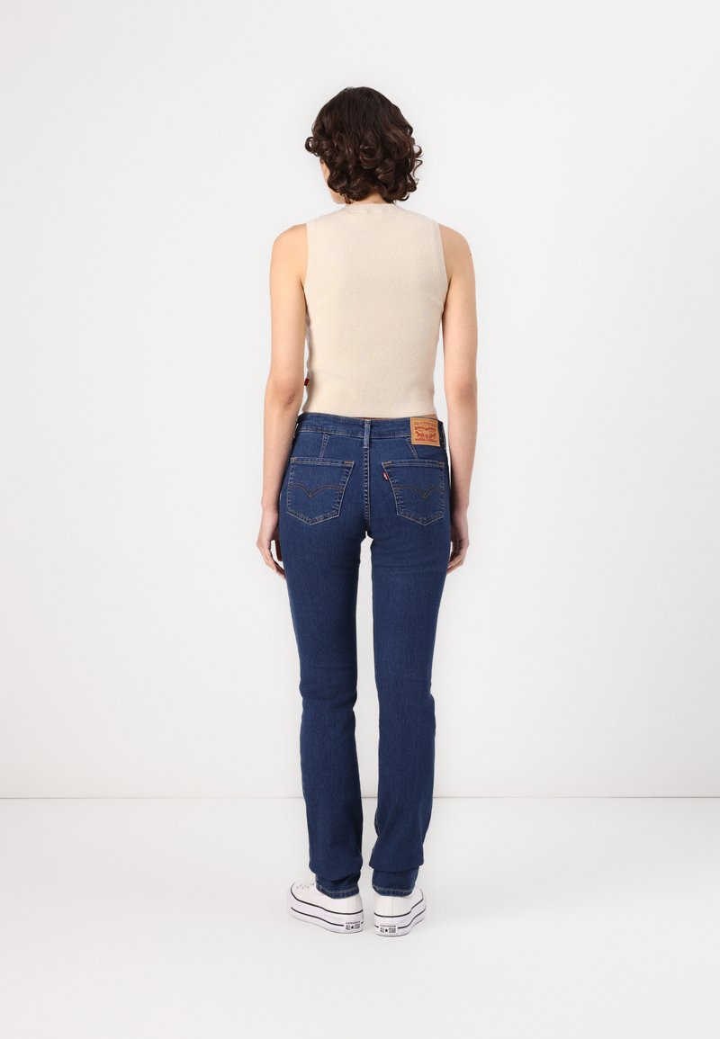 Vaqueros de denim de color azul oscuro, diseño de pierna recta, con un ligero estiramiento y costuras características en los bolsillos traseros; combinados con una blusa sin mangas de color beige.