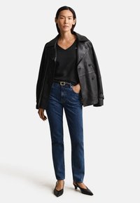 Γυναίκα που φοράει μαύρο δερμάτινο μπουφάν πάνω από μαύρο τοπ με V-λαιμόκοψη, μπλε τζιν με ζώνη και μαύρα μυτερά τακούνια, στέκεται με το ένα χέρι στην τσέπη.