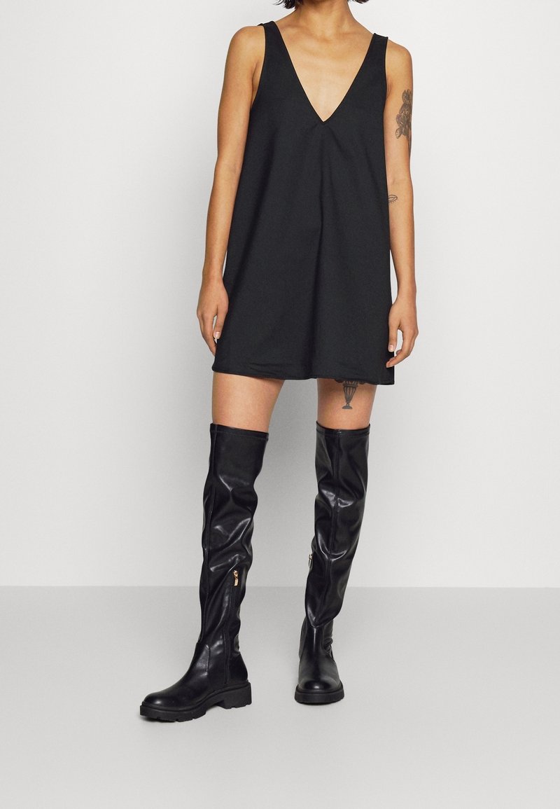 Schwarzes ärmelloses Kleid mit V-Ausschnitt, das ein fließendes Design aufweist. Kombiniert mit kniehohen, glänzenden schwarzen Lederstiefeln mit Reißverschlüssen.