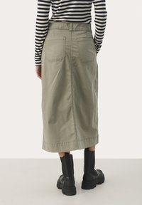 Jupe midi en coton vert avec deux poches arrière, une coupe droite et un ourlet légèrement évasé. Associée à des bottines noires.