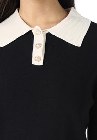 Czarny prążkowany top z kontrastującym, białym kołnierzykiem. Posiada cztery guziki ozdobione perłowymi akcentami. Zbliżenie na linię dekoltu.