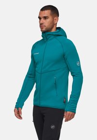 Giacca in pile turchese con cappuccio, tasche laterali con zip e polsini elastici. Presenta cuciture a contrasto e un logo sul petto e sulla manica.
