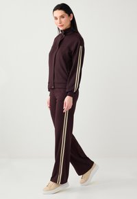 Survêtement bordeaux avec une veste zippée et un pantalon à jambes larges, présentant des rayures verticales crème sur les côtés. Fabriqué en tissu doux et lisse.