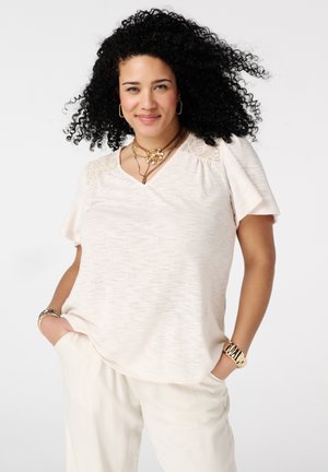 Vrouw met krullend haar die een lichtbeige top met korte mouwen en kanten details draagt, lichte broek en gouden sieraden, glimlachend met haar handen in haar zakken.