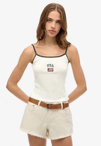 Weißes, geripptes Tanktop mit marineblauen Akzenten, versehen mit "USA"-Stickerei und einem kleinen Flaggenaufnäher; kombiniert mit hochgeschnittenen beigen Shorts und einem braunen Gürtel.