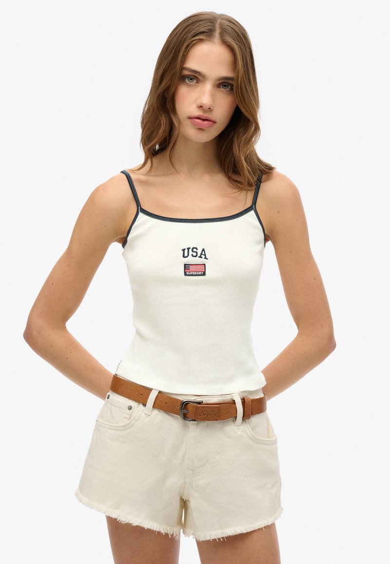Weißes, geripptes Tanktop mit marineblauen Akzenten, versehen mit "USA"-Stickerei und einem kleinen Flaggenaufnäher; kombiniert mit hochgeschnittenen beigen Shorts und einem braunen Gürtel.